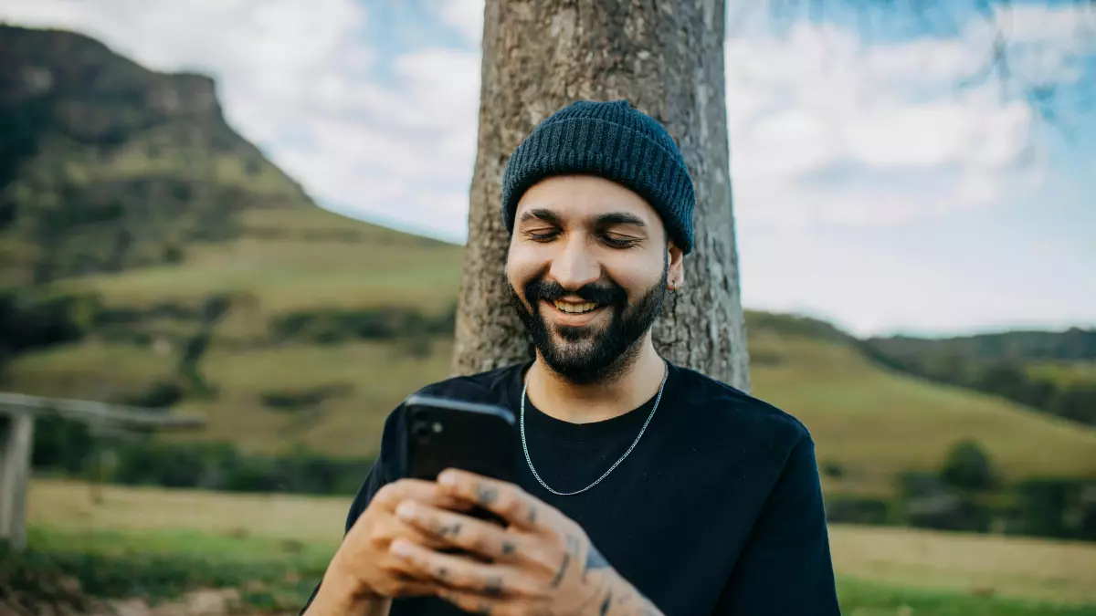 A young man with a beard and a black beanie is smiling while looking at his phone. He is standing in front of a tree with a green hill and blue sky in the background.